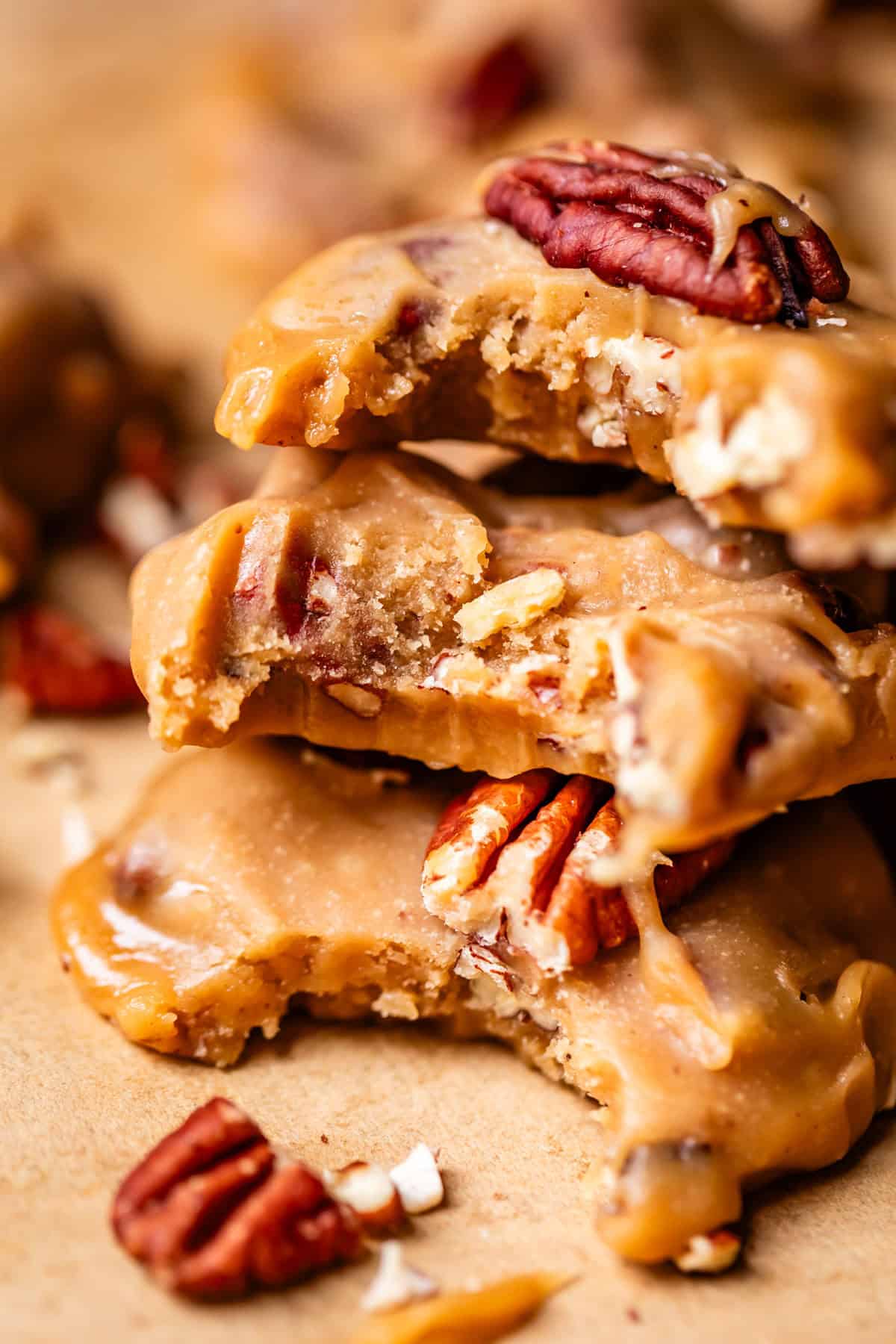 Close-up of stacked pecan pralines with a creamy caramel coating, showing a chewy texture and whole pecans. A few pralines and pecan pieces are scattered on a brown surface.