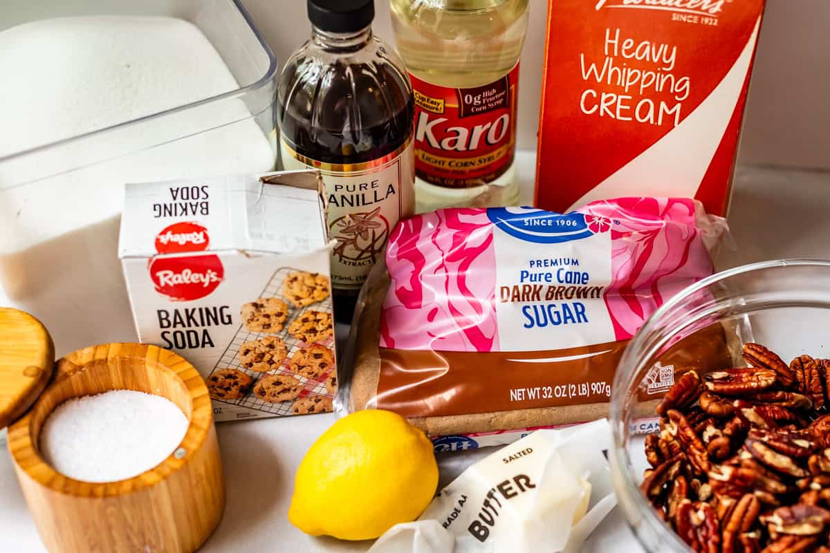 Various baking ingredients are arranged on a counter, including brown sugar, heavy whipping cream, vanilla extract, corn syrup, baking soda, salt, a lemon, butter, pecans, and a container of white sugar.