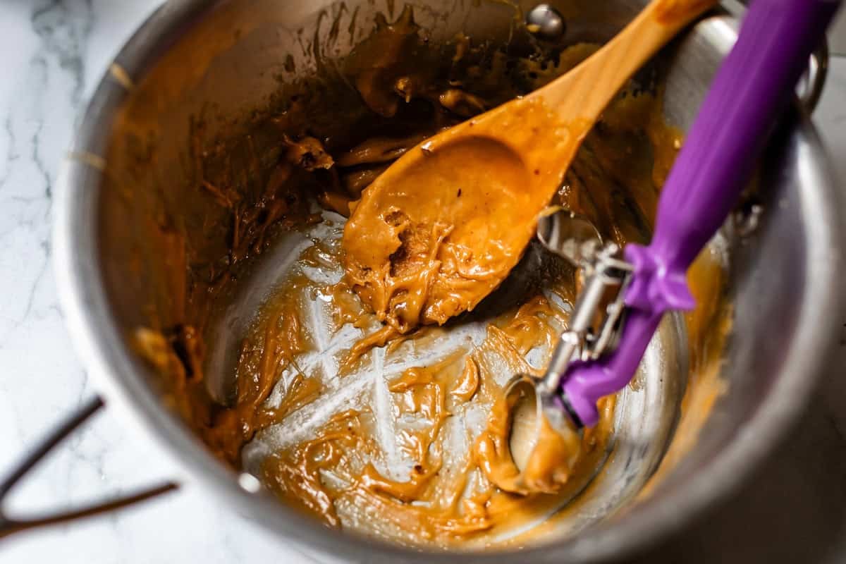 A metal mixing bowl with remnants of thick, light brown batter, a wooden spoon, and a purple-handled cookie scoop, all resting on a white marble surface.
