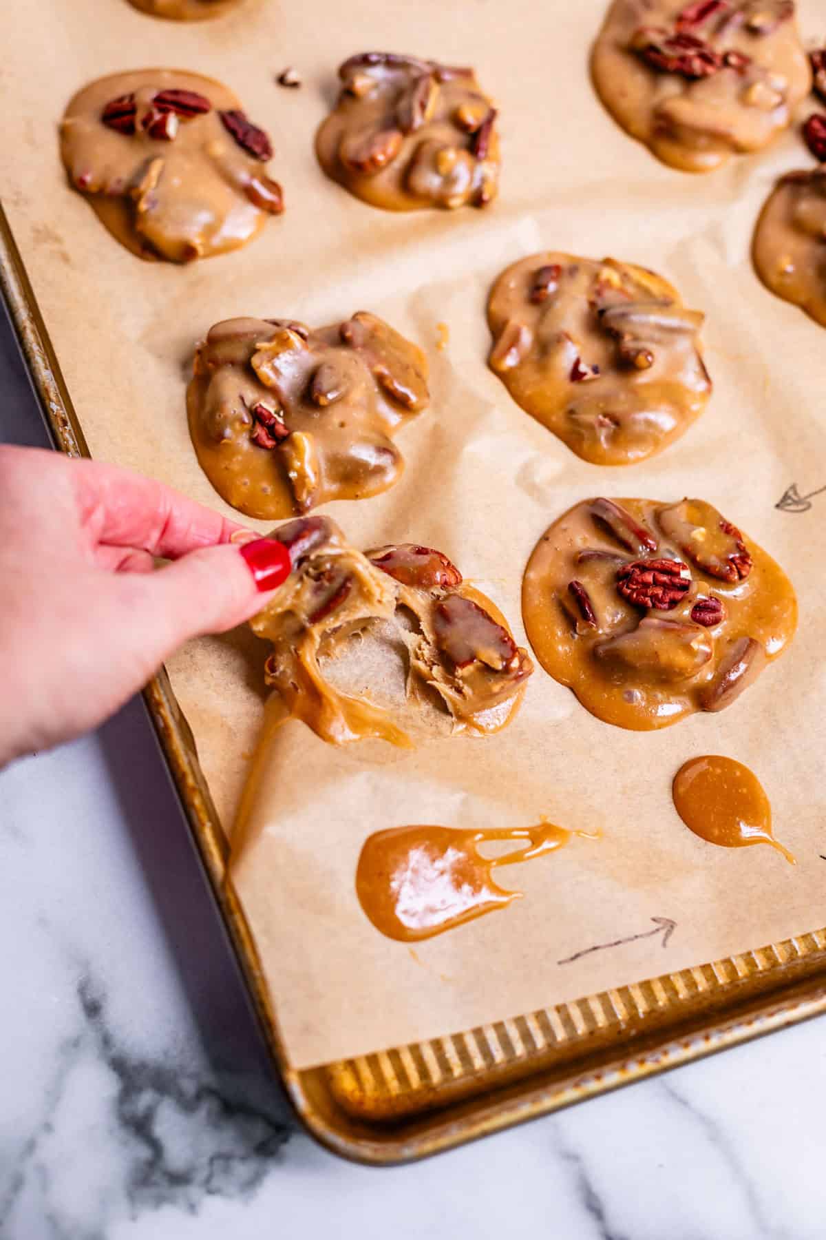 A hand lifts a pecan praline cookie from a baking sheet lined with parchment paper. Several other pralines with glossy, caramel coating and pecan pieces are visible on the sheet.