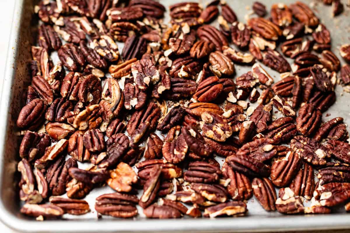 A baking tray filled with roasted pecan halves and pieces, spread out in a single layer. The nuts appear toasted and slightly seasoned.