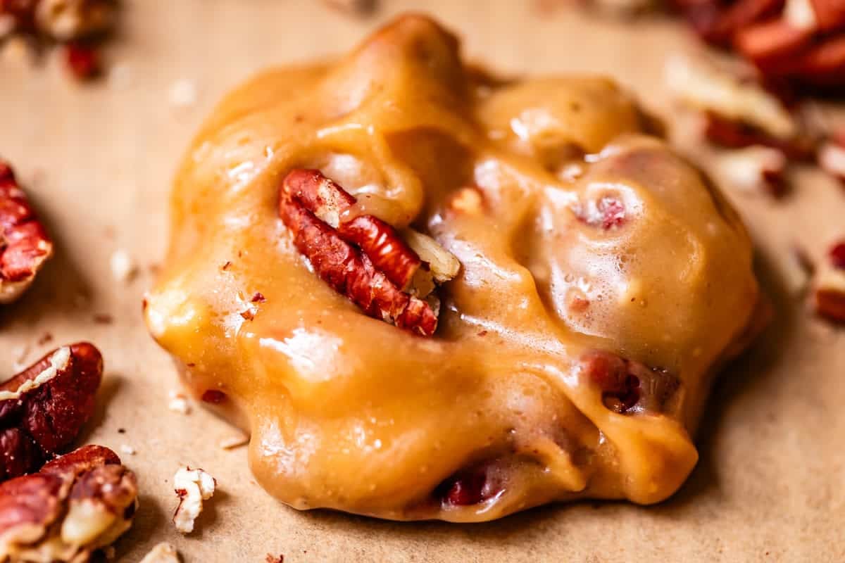 A close-up of a golden brown pecan praline on parchment paper, with whole and chopped pecans visible in the creamy, glossy candy. Scattered pecan pieces surround the praline.
