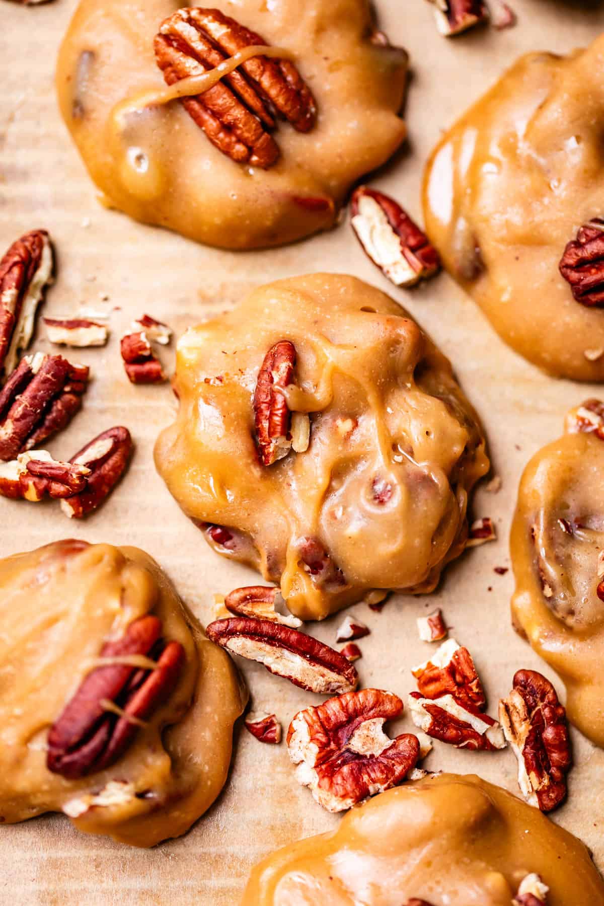 Close-up of creamy praline cookies topped and surrounded by whole and chopped pecans, displayed on parchment paper. The cookies have a glossy, caramel-like texture.