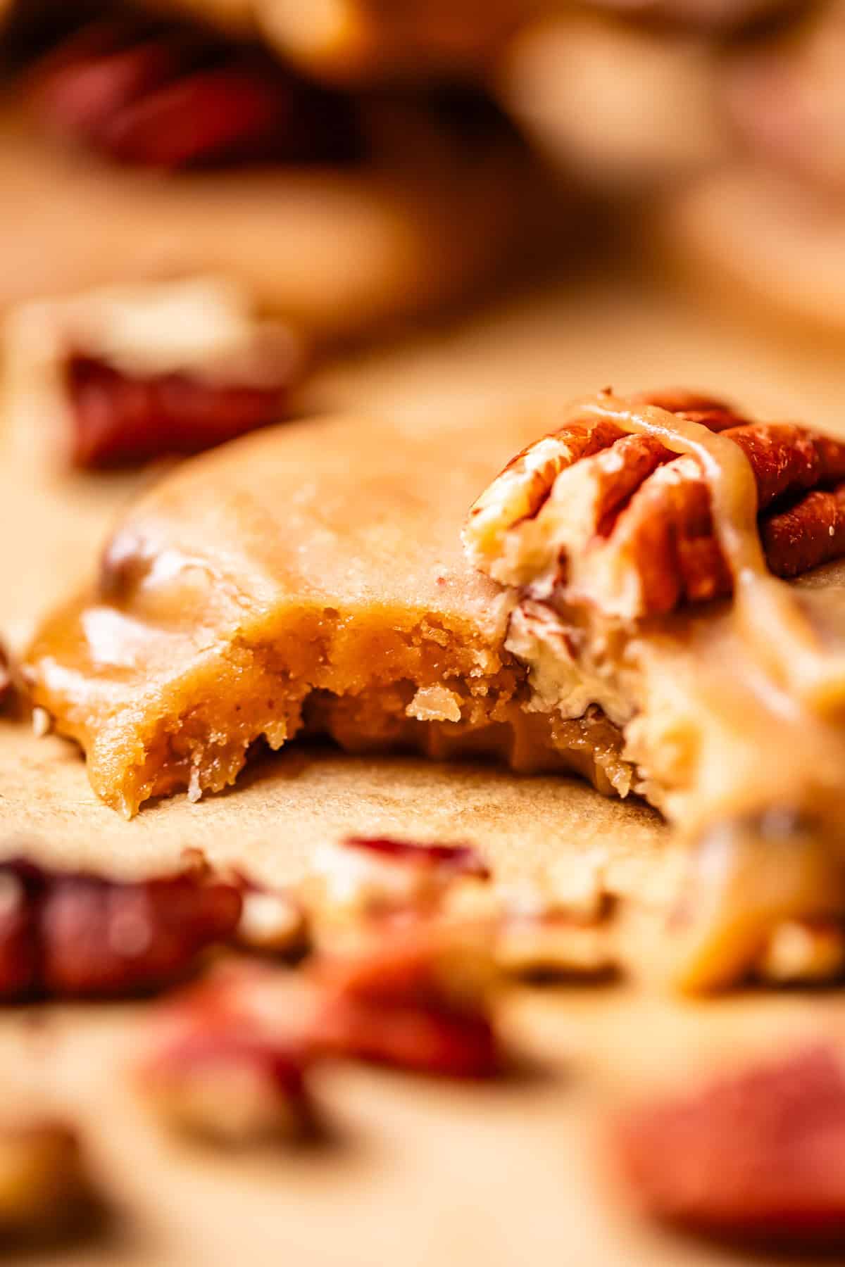 A close-up of a praline cookie topped with pecans, with a bite taken out, showing its soft interior. Crumbled pecan pieces are scattered around on a light-colored surface.