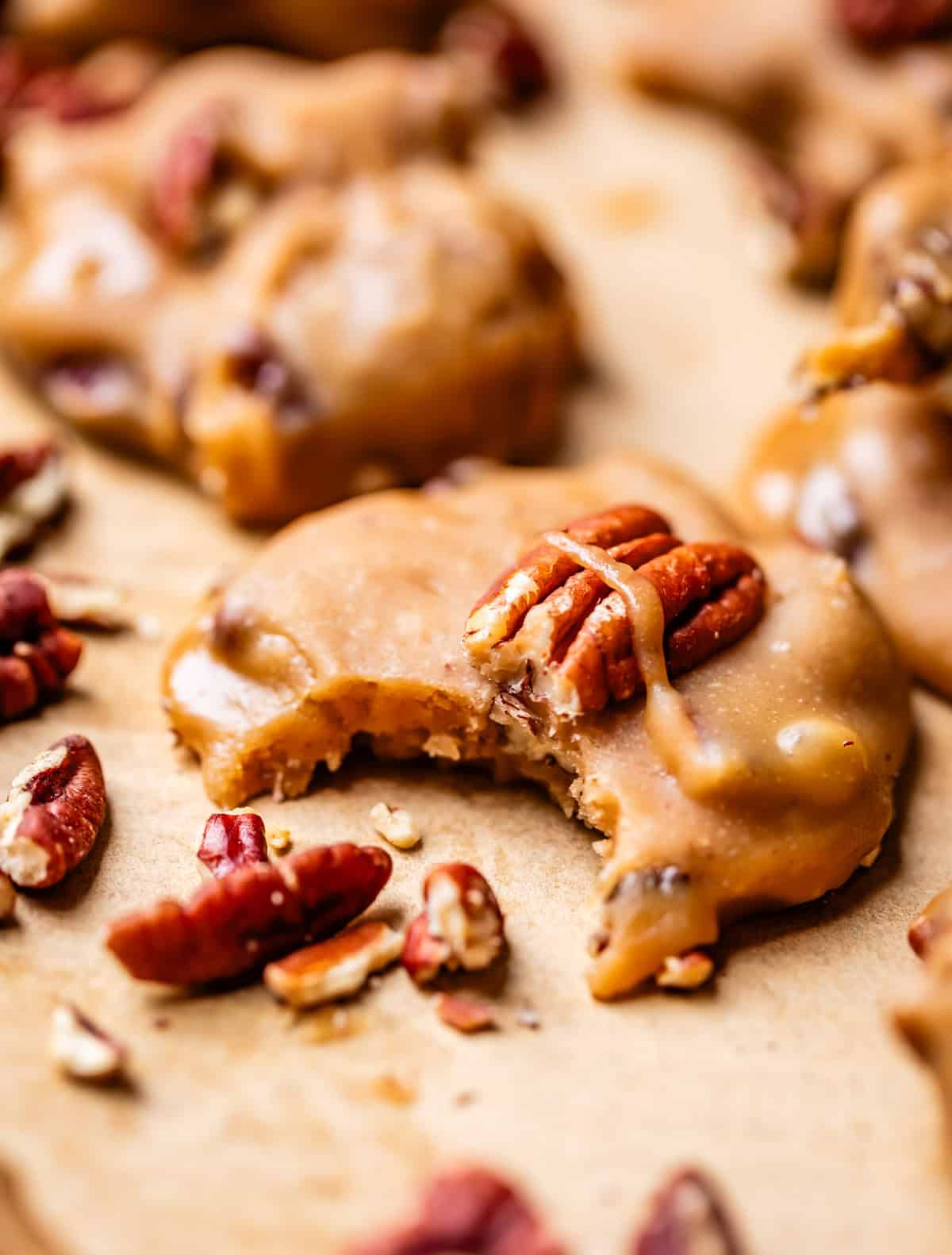 A close-up of pecan pralines on parchment paper, with one praline missing a bite. Whole and chopped pecans are scattered around the creamy, caramel-colored candies.