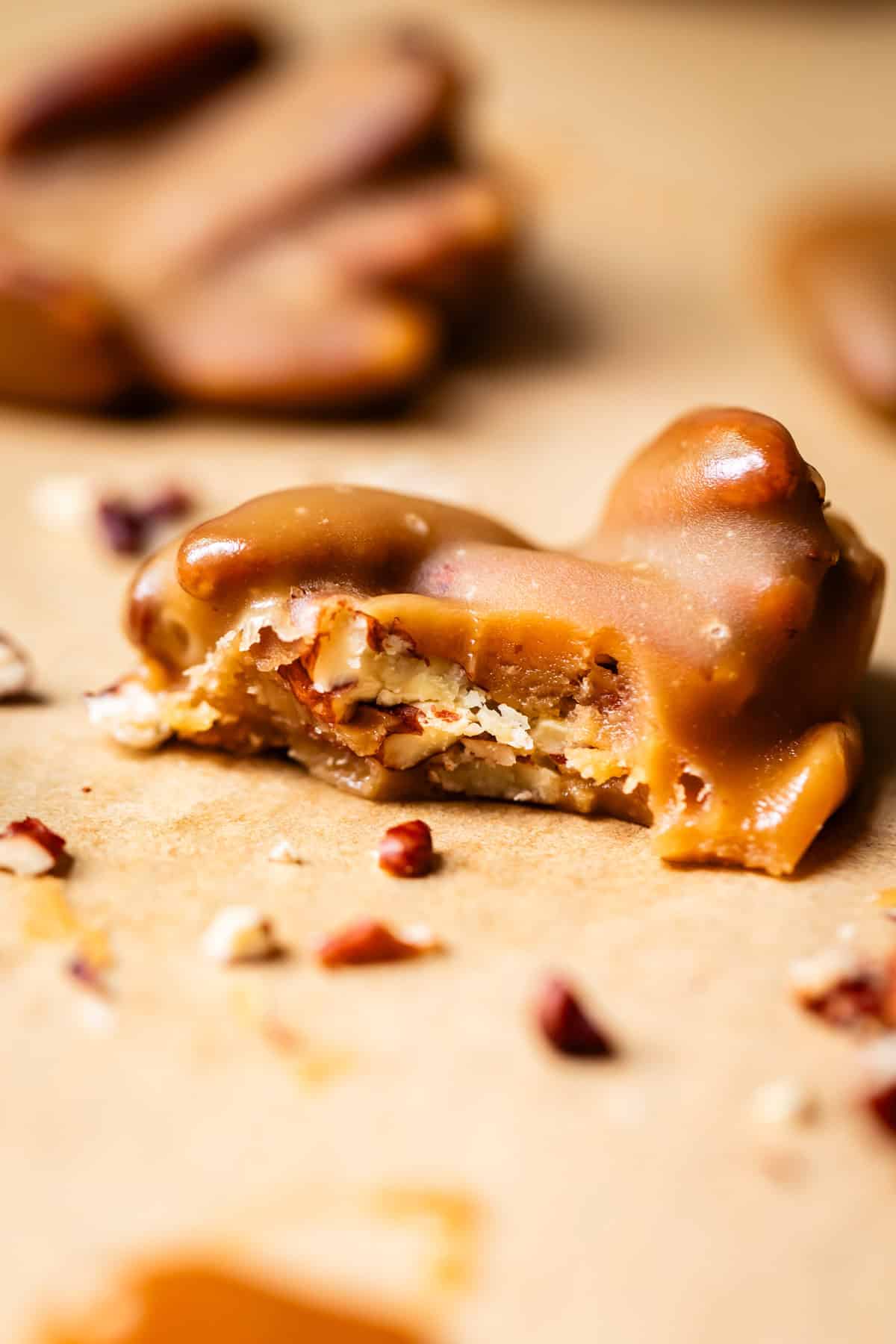 A close-up of a pecan praline candy with a bite taken out, showing crunchy pecans and creamy caramel. Crumbled pecan pieces are scattered on a light, textured surface around the candy.