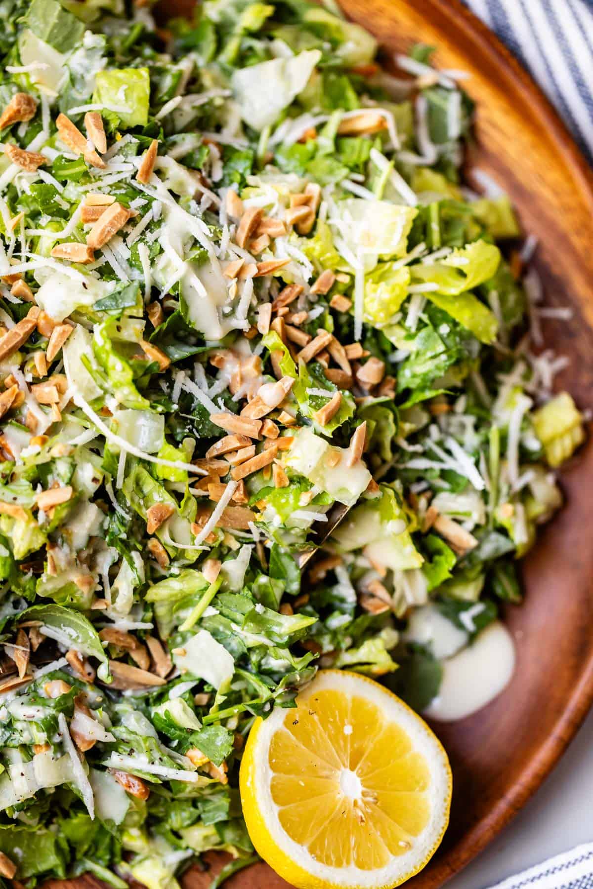 A close-up of a fresh green salad on a wooden plate, topped with shredded cheese, slivered almonds, and creamy dressing, with a half lemon placed on the side.