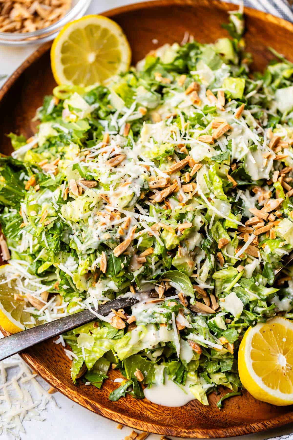 A wooden bowl filled with chopped green salad, shredded cheese, sunflower seeds, creamy dressing, and lemon wedges. A serving spoon rests in the bowl, and a small dish of sunflower seeds is in the background.