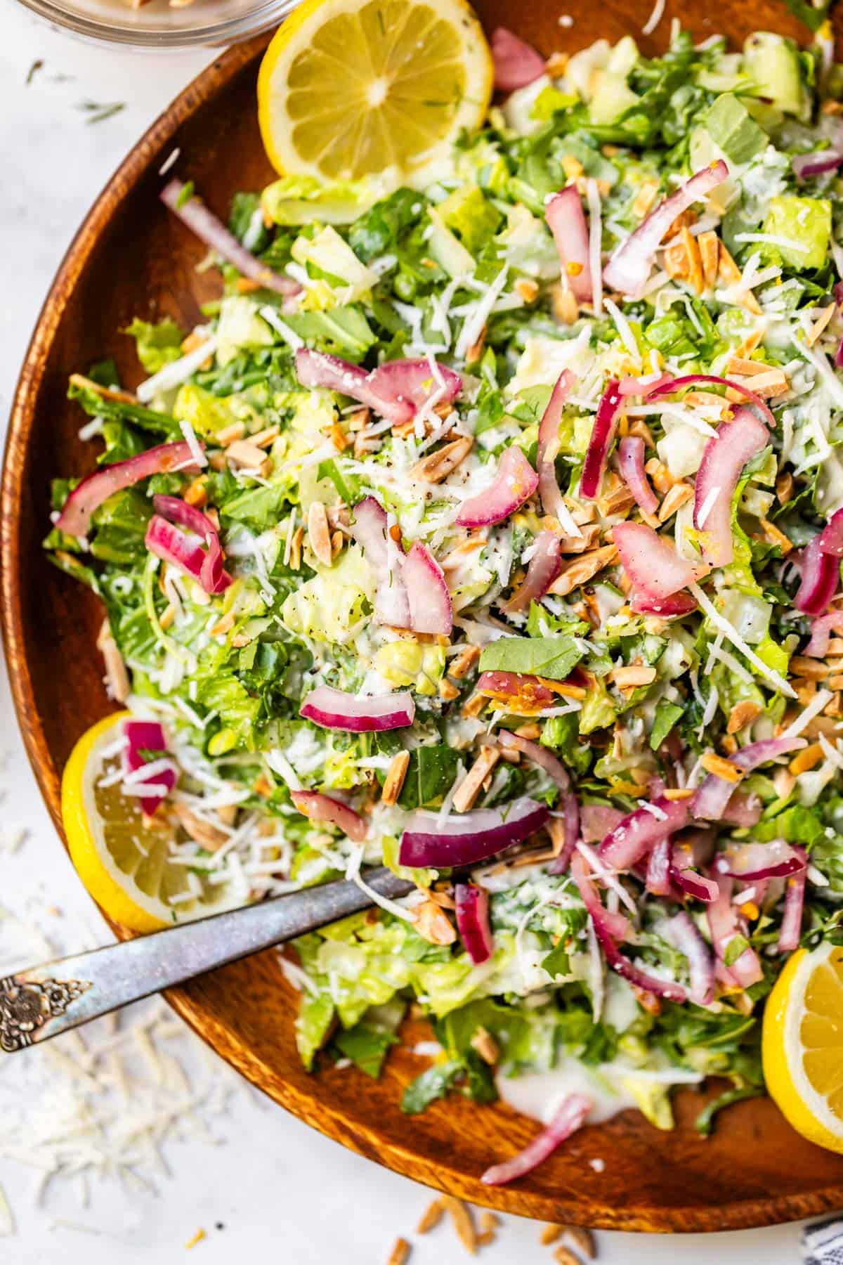 A wooden bowl filled with chopped salad topped with shredded cheese, sliced red onions, slivered almonds, and creamy dressing, garnished with lemon slices, with a silver serving spoon inside.