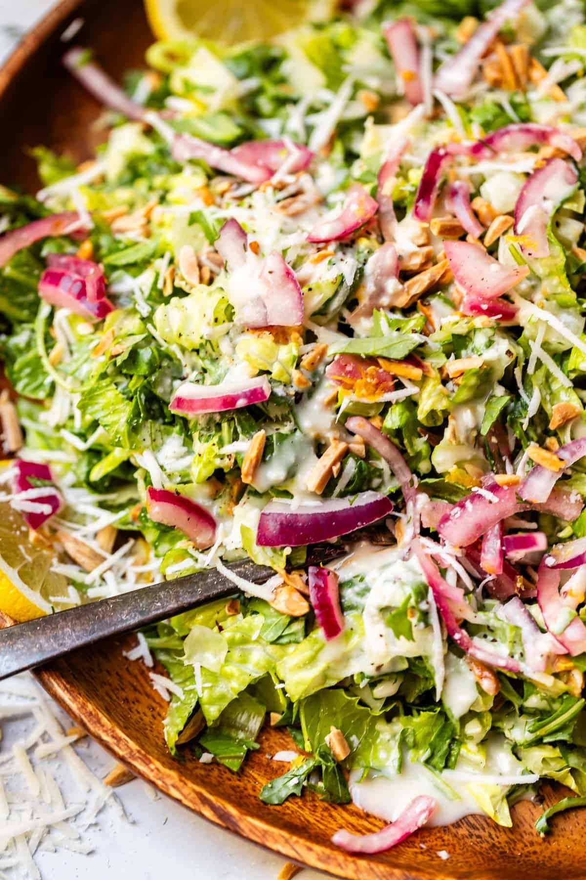 A close-up of a fresh green salad with chopped lettuce, pickled red onions, shredded cheese, sunflower seeds, and creamy dressing, served in a wooden bowl with lemon wedges on the side.