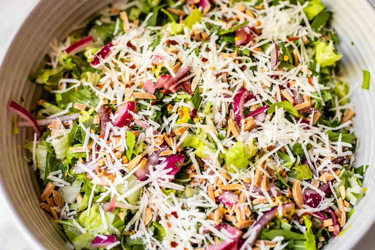 A close-up of a fresh salad with chopped romaine lettuce, shredded Parmesan cheese, sliced red onions, toasted almonds, and green herbs in a bowl.