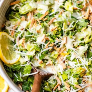 A close-up of a fresh green salad with shredded lettuce, grated cheese, slivered almonds, and lemon slices, drizzled with creamy dressing and served in a bowl with a wooden spoon.