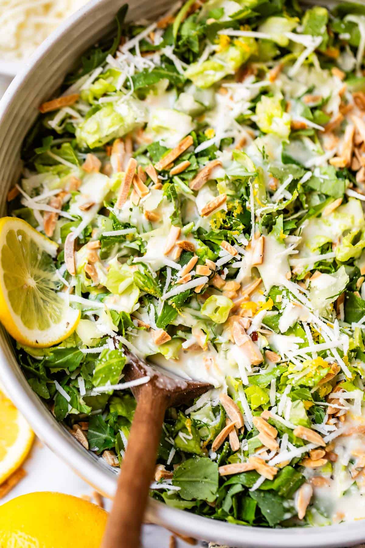 A close-up of a fresh green salad with shredded lettuce, grated cheese, slivered almonds, and lemon slices, drizzled with creamy dressing and served in a bowl with a wooden spoon.