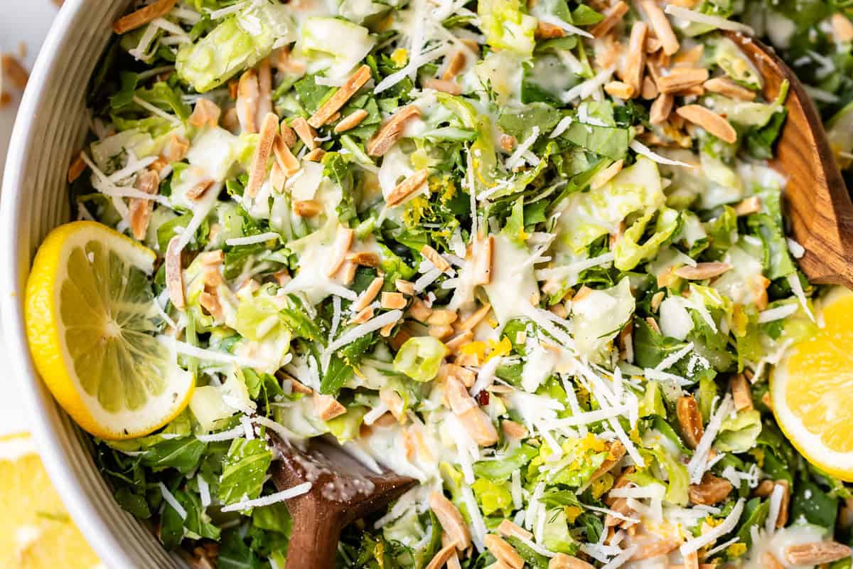 A close-up of a fresh salad with chopped greens, shredded cheese, toasted almonds, creamy dressing, and lemon slices, being mixed with wooden salad servers.