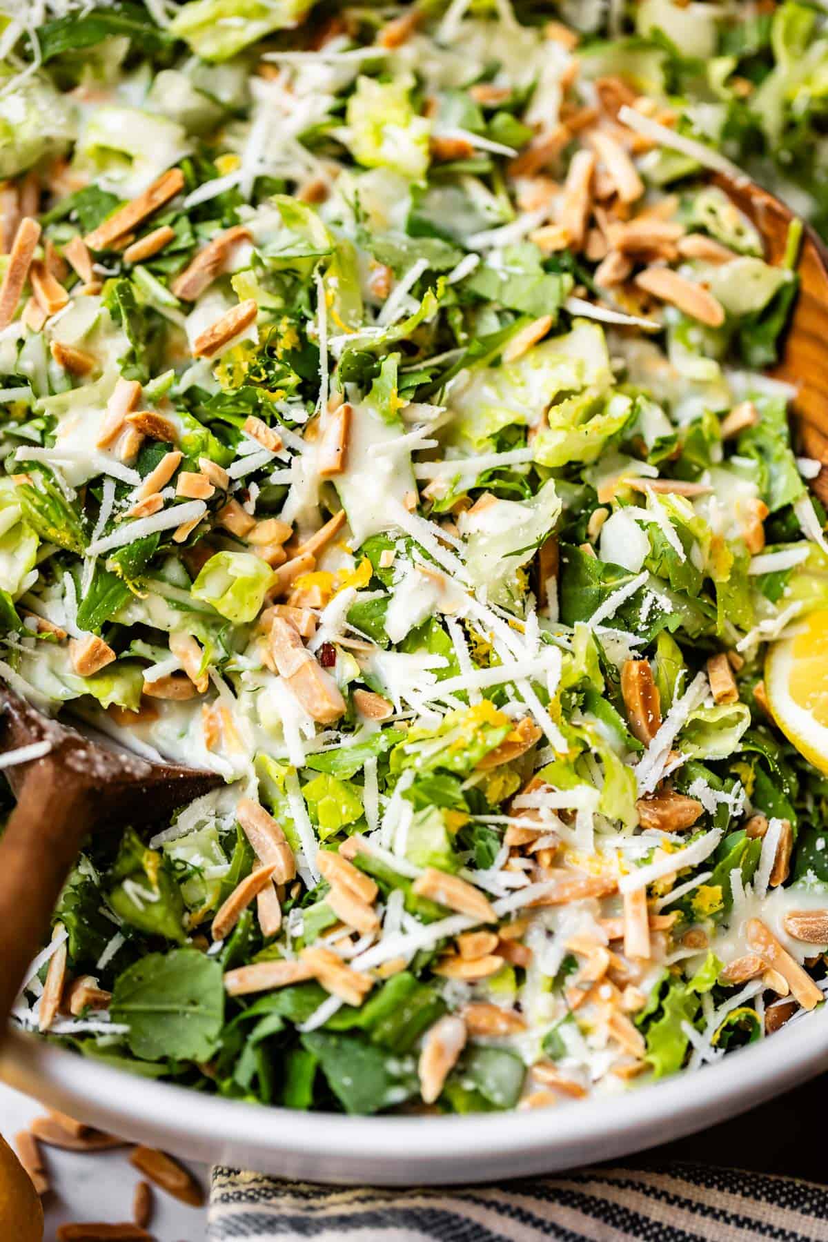A close-up of a fresh green salad with chopped lettuce, shredded cheese, slivered almonds, and creamy dressing, garnished with a lemon wedge.