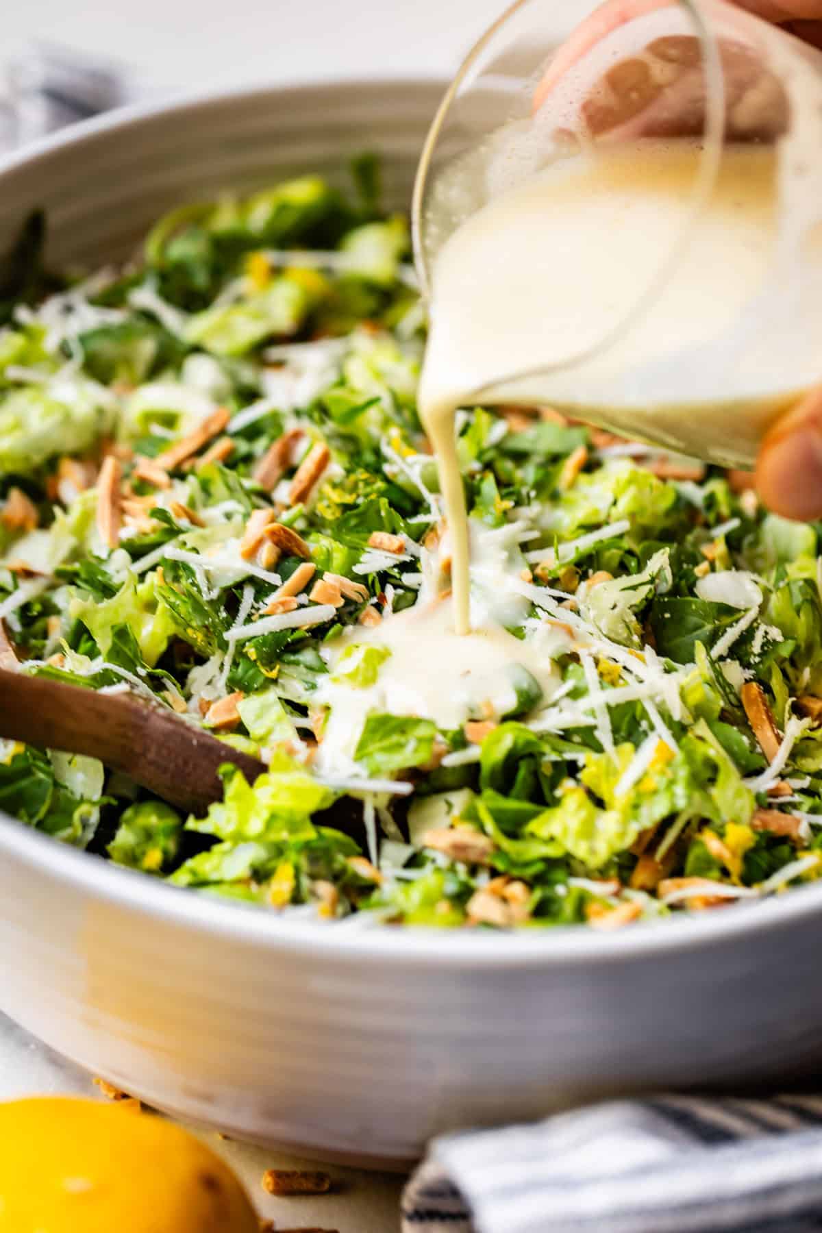 A close-up of a bowl of chopped green salad with shredded cheese and nuts, as creamy dressing is being poured over it from a small glass container. A wooden spoon is visible in the salad.
