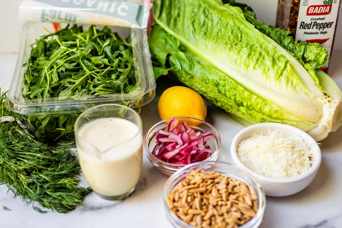 Fresh salad ingredients including arugula, romaine lettuce, a lemon, fresh dill, a jar of red pepper flakes, sliced red onions, shredded cheese, slivered almonds, and a cup of creamy salad dressing on a white surface.