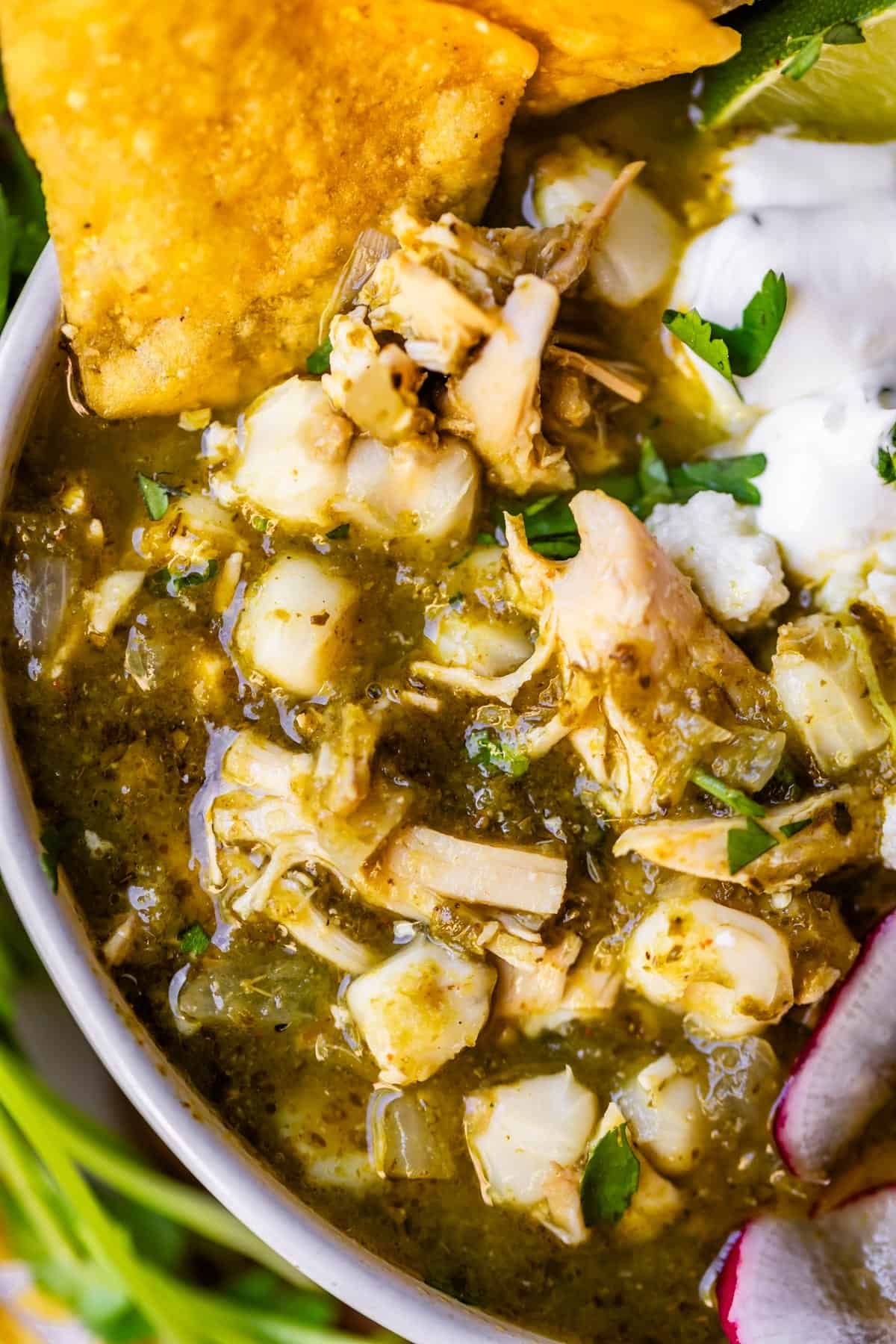 hero?? A close-up of a bowl of green chicken pozole, featuring shredded chicken, hominy, tortilla chips, fresh cilantro, sour cream, a lime wedge, and sliced radish. The soup has a rich green broth.
