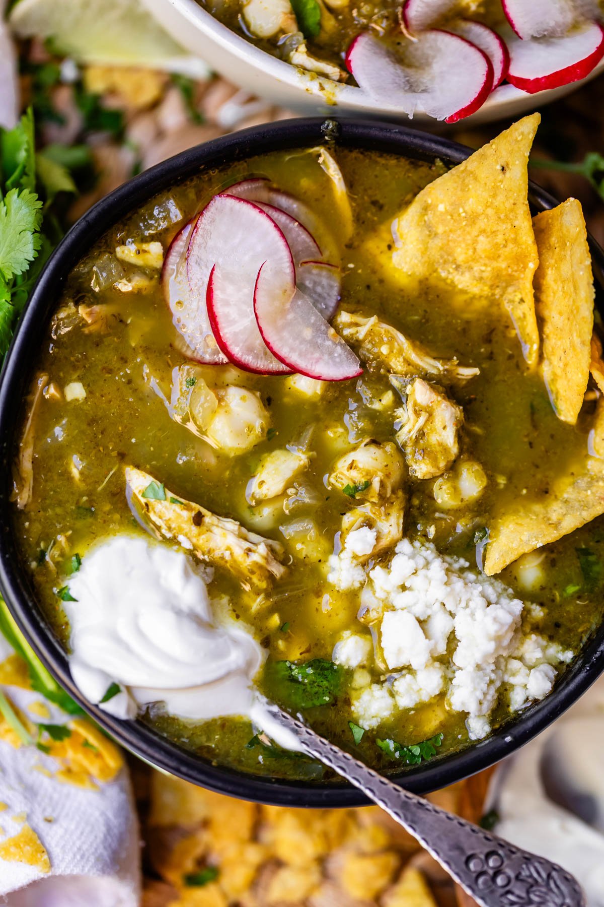 A bowl of green chicken pozole topped with sliced radishes, crumbled cheese, tortilla chips, and a dollop of sour cream, with a spoon resting inside the bowl.