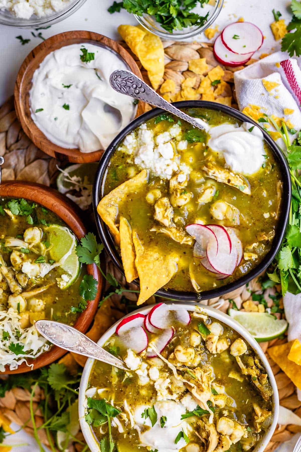 Bowls of green chicken pozole topped with radish slices, tortilla chips, sour cream, and fresh cilantro, surrounded by lime wedges, corn chips, and additional garnishes on a textured surface.