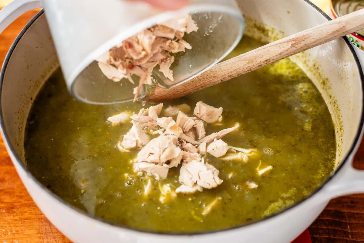 A hand pours cooked, shredded chicken from a bowl into a pot of green soup with a wooden spoon inside, preparing a homemade meal.