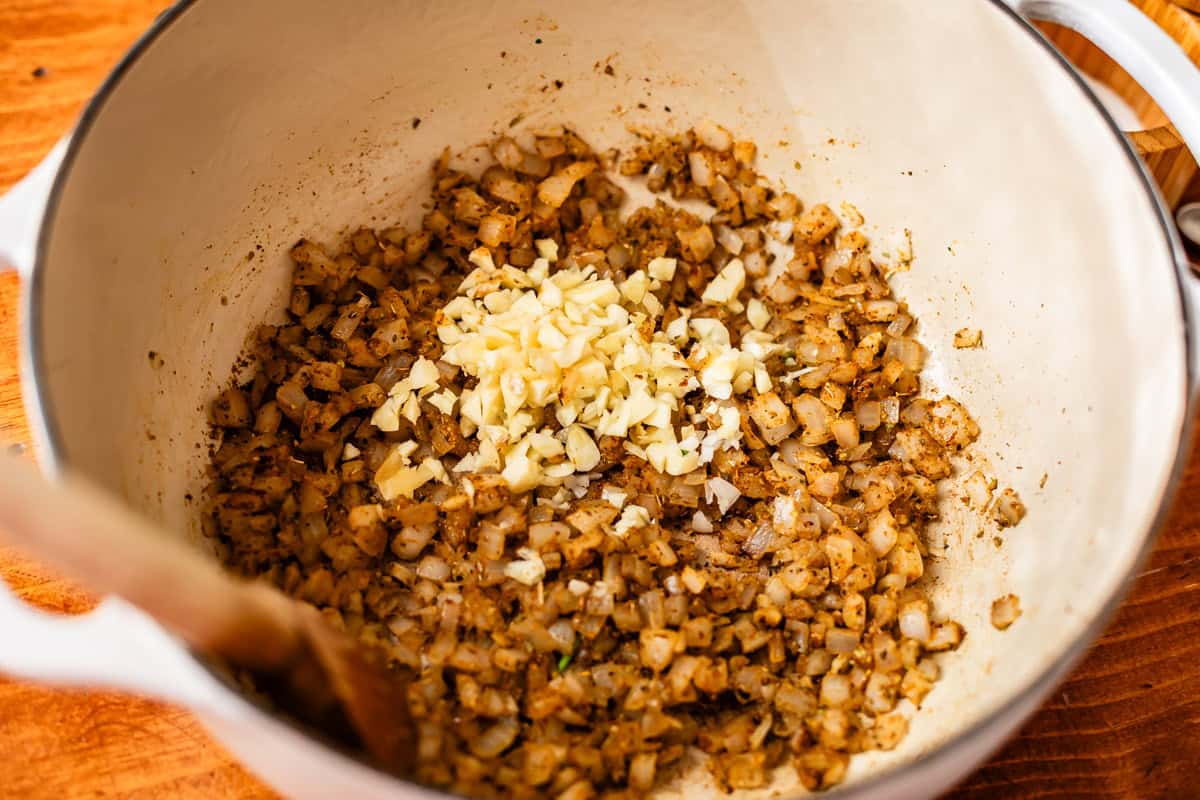 Chopped onions and minced garlic are being saut&eacute;ed with spices in a white pot, stirred with a wooden spoon, on a wooden surface.