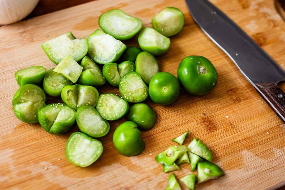 Chopped and whole green tomatillos on a wooden cutting board next to a kitchen knife.