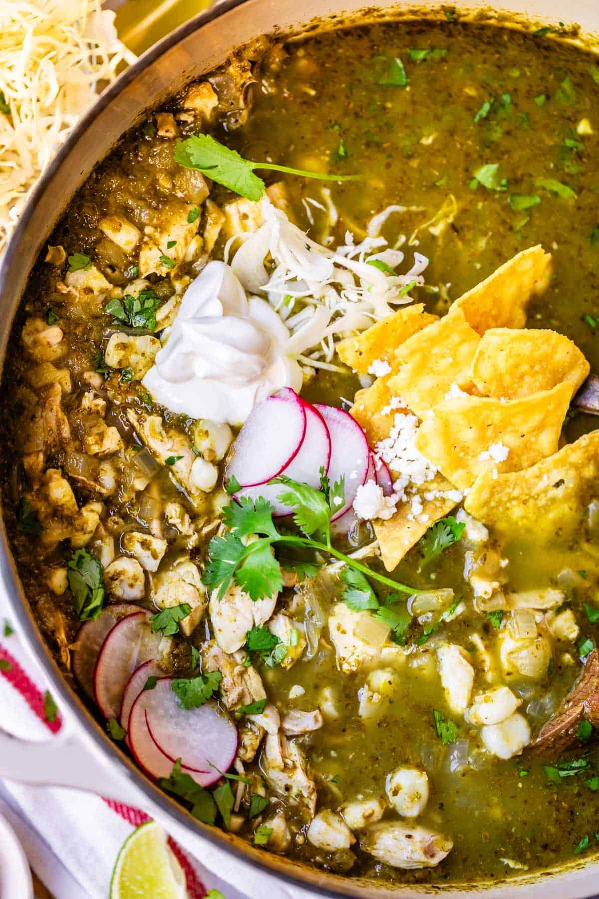 A bowl of green pozole soup garnished with sliced radishes, fresh cilantro, tortilla chips, shredded cheese, and a dollop of sour cream, with visible pieces of hominy and chicken in a vibrant green broth.