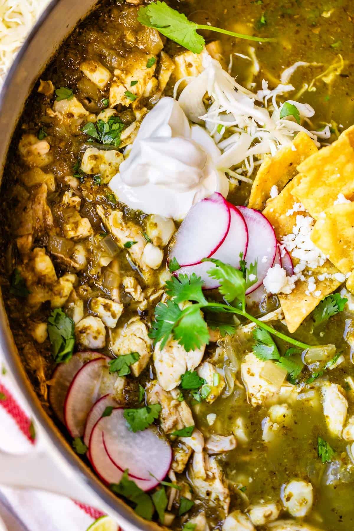 A close-up of a bowl of green pozole topped with sliced radishes, shredded cabbage, tortilla chips, fresh cilantro, a dollop of sour cream, and crumbled cheese. The soup contains hominy and chunks of chicken.