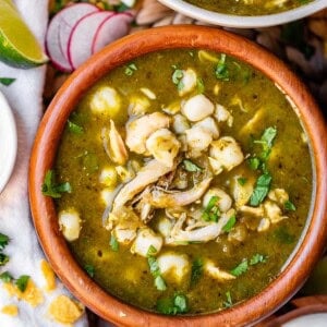 A wooden bowl filled with green pozole soup, topped with shredded chicken, hominy, and chopped cilantro. Surrounding the bowl are lime wedges, sliced radishes, tortilla chips, and a small bowl of sour cream.