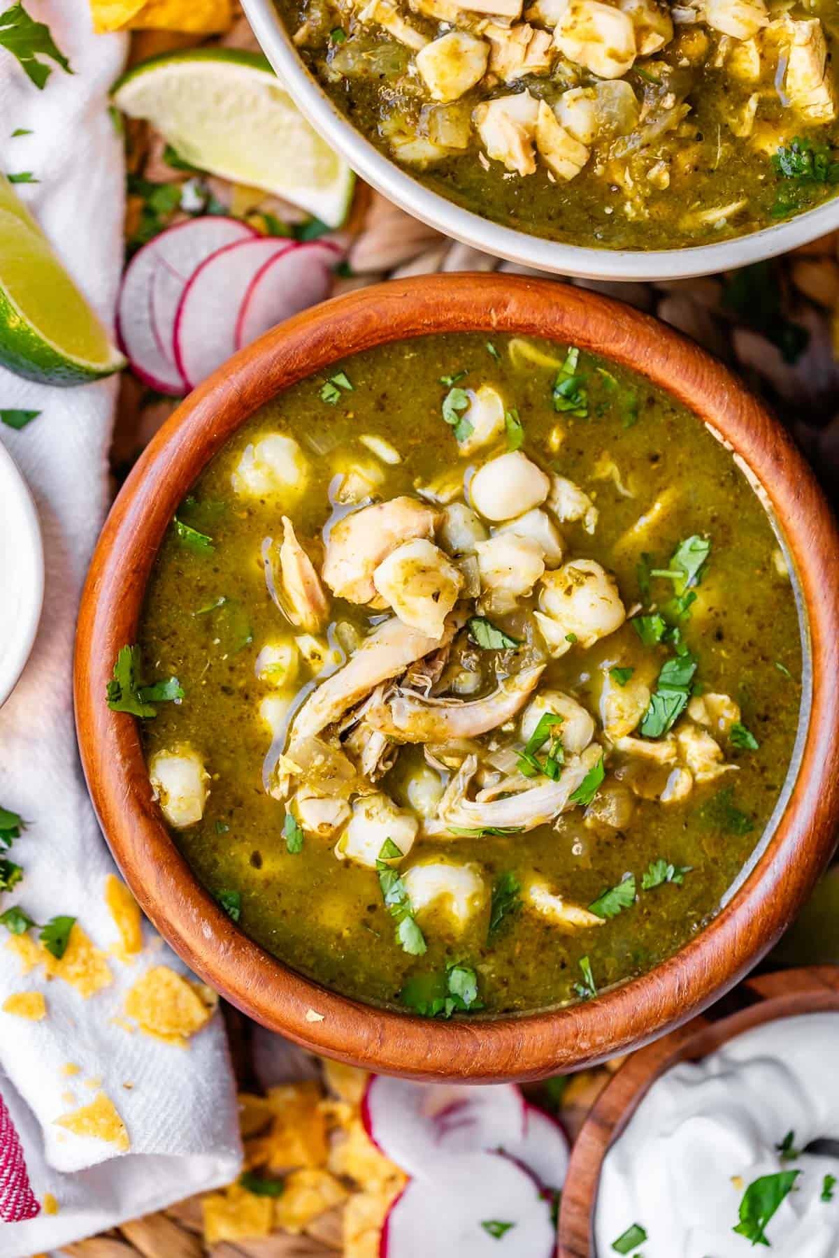 A wooden bowl filled with green pozole soup, topped with shredded chicken, hominy, and chopped cilantro. Surrounding the bowl are lime wedges, sliced radishes, tortilla chips, and a small bowl of sour cream.