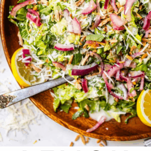 A wooden bowl filled with lemon arugula salad, topped with grated cheese, sliced red onions, seeds, and lemon wedges. A fork rests in the bowl. Text at the bottom reads “Lemon Arugula Salad.”.