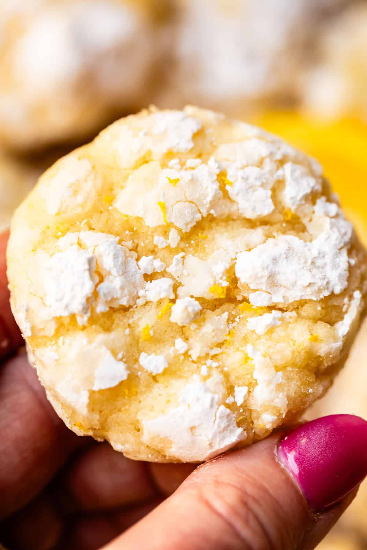 A hand with pink nail polish holds a round lemon crinkle cookie topped with powdered sugar and yellow cracks.