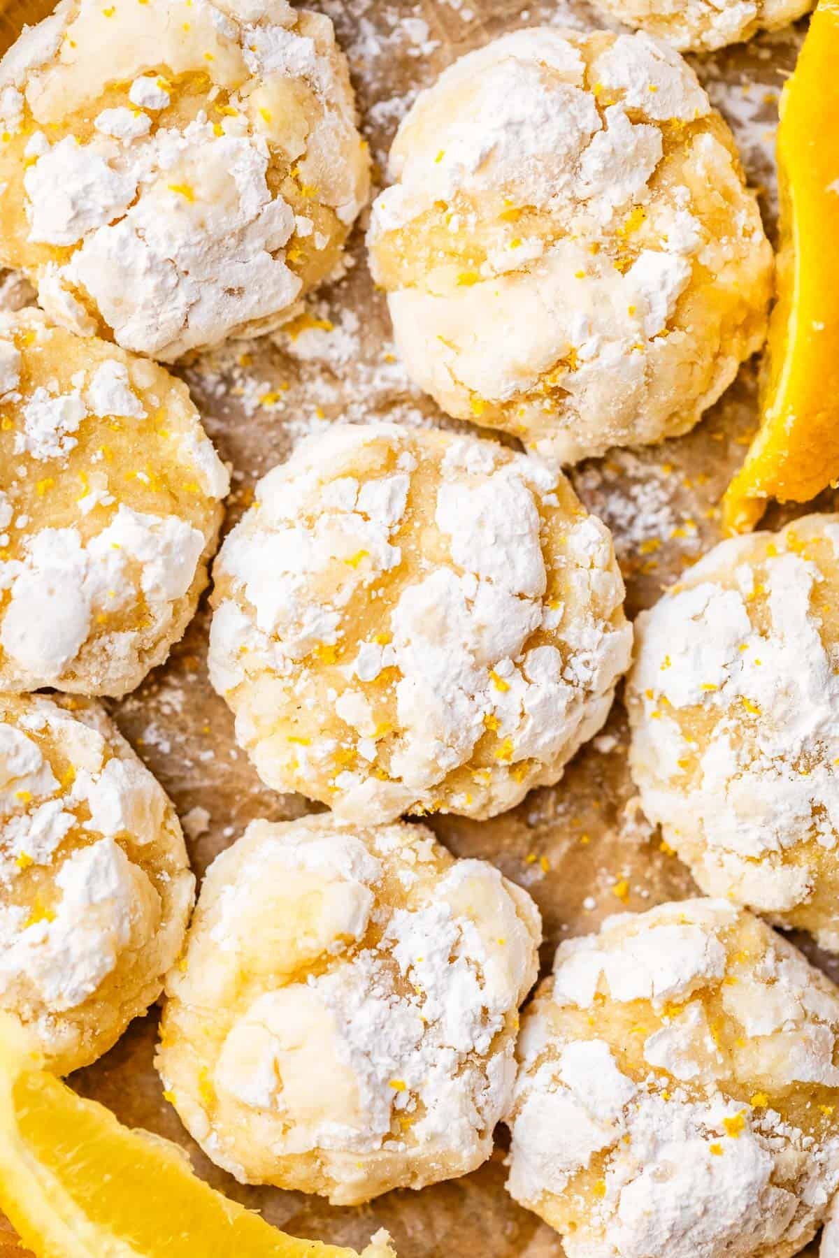 Close-up of several lemon crinkle cookies coated in powdered sugar, arranged on parchment paper with a few orange slices visible at the edges. The cookies are golden yellow and have a cracked, textured surface.
