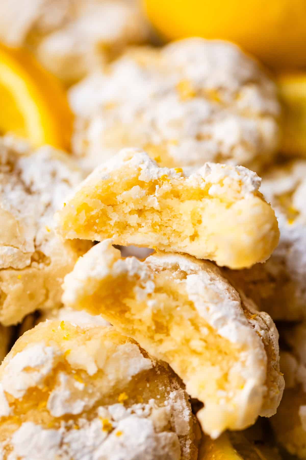 A close-up of soft, lemon crinkle cookies dusted with powdered sugar. One cookie is broken in half, showing its moist, yellow interior, with more cookies and lemon slices in the background.