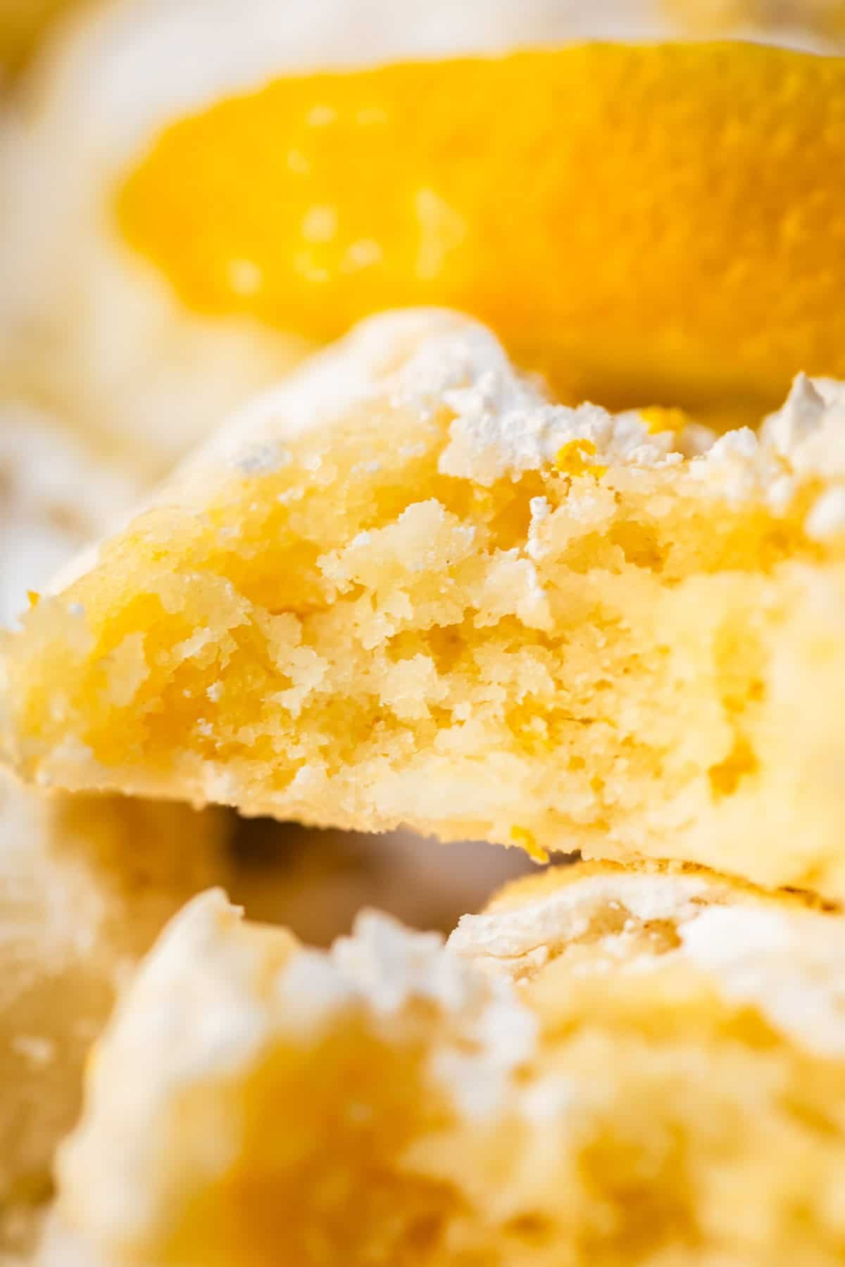 A close-up of a soft, crumbly lemon cookie dusted with powdered sugar, with a slice of lemon in the background.