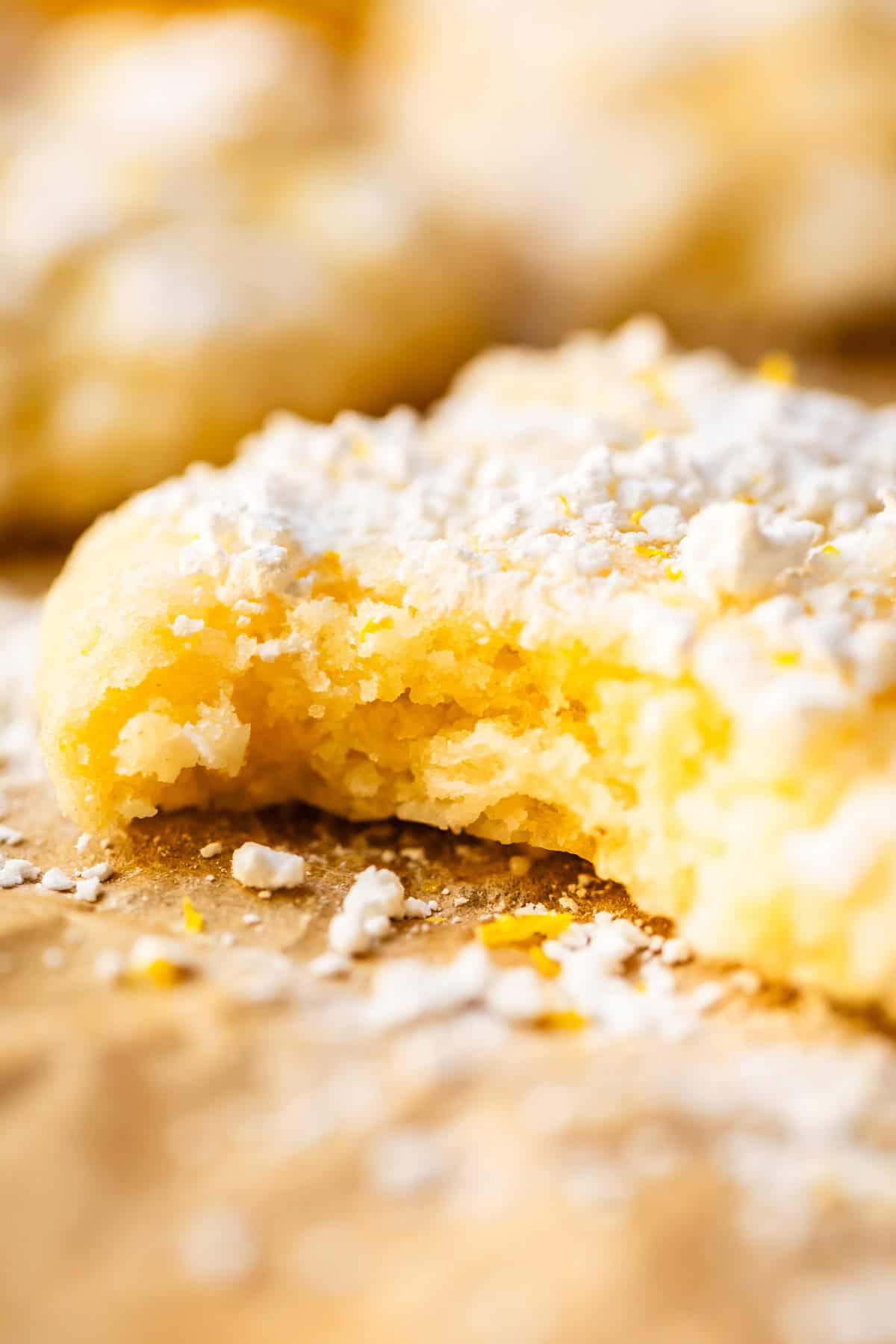 A soft, yellow cookie dusted with powdered sugar and a few lemon zest flakes, shown in close-up with a bite taken out of it. The background is blurred, highlighting the cookies texture.