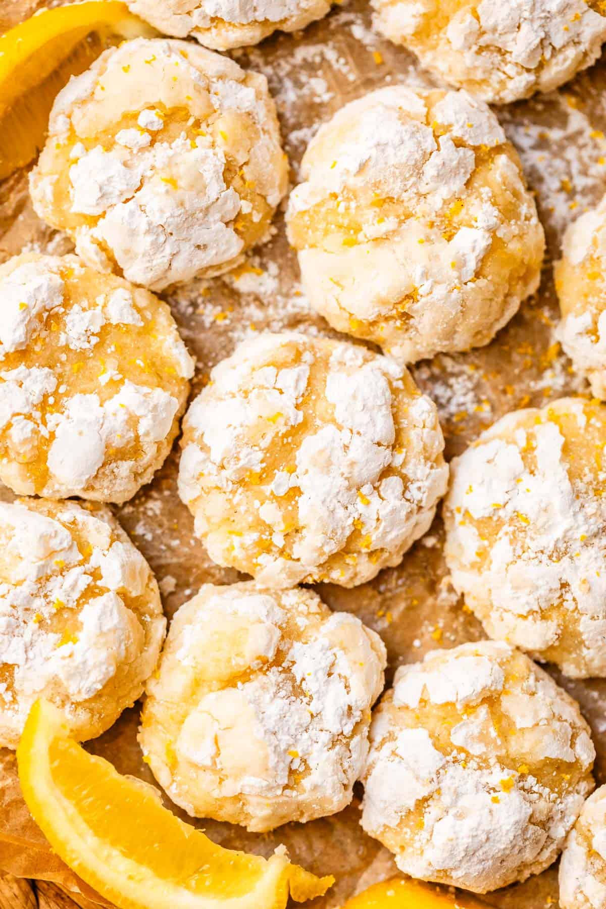 Close-up of several orange crinkle cookies coated in powdered sugar, arranged on parchment paper with a few orange wedges scattered around. The cookies have a cracked, textured surface and a light, fluffy appearance.