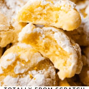 A close-up of soft, powdered sugar-covered lemon crinkle cookies stacked on top of each other, with a slice of lemon and the text “Totally from scratch Lemon Crinkle Cookies” at the bottom.