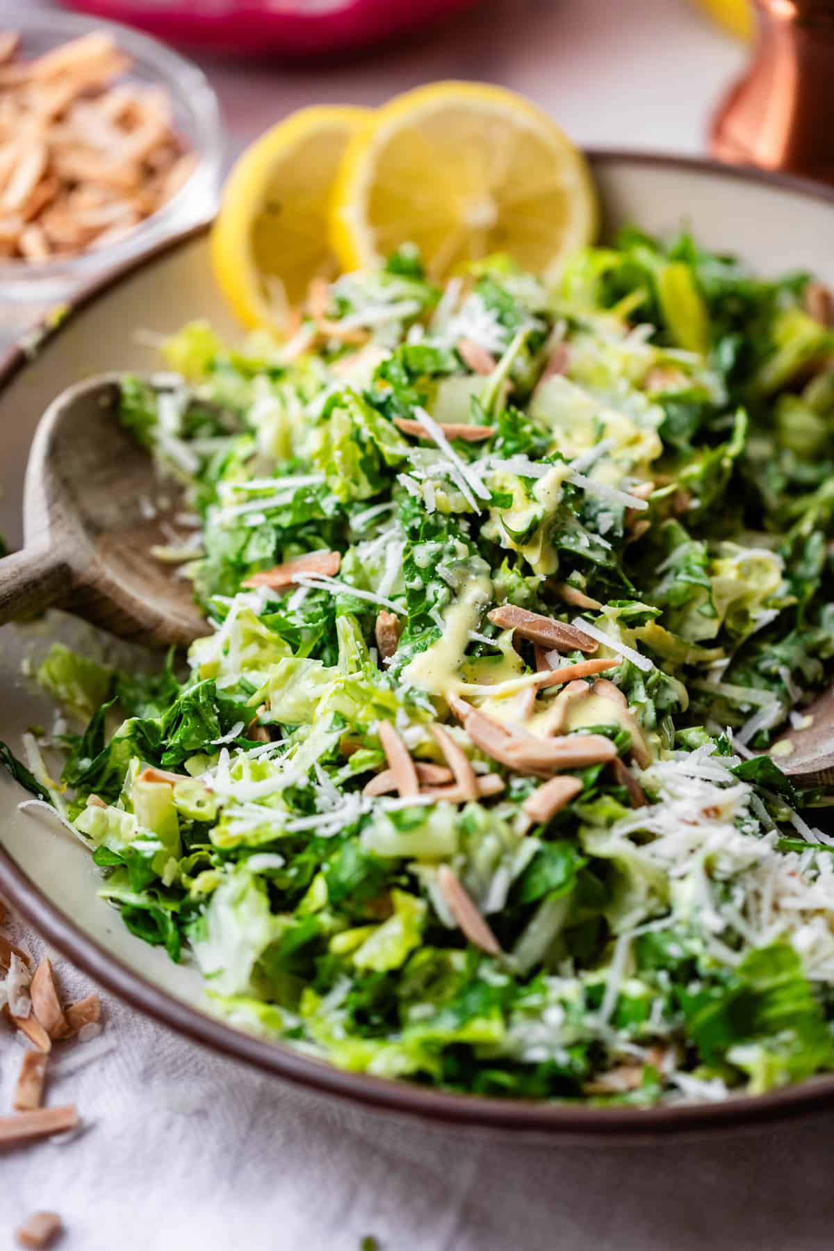 A close-up of a fresh green salad with shredded lettuce, grated cheese, slivered almonds, and creamy dressing, served in a bowl with lemon slices and wooden salad tongs.