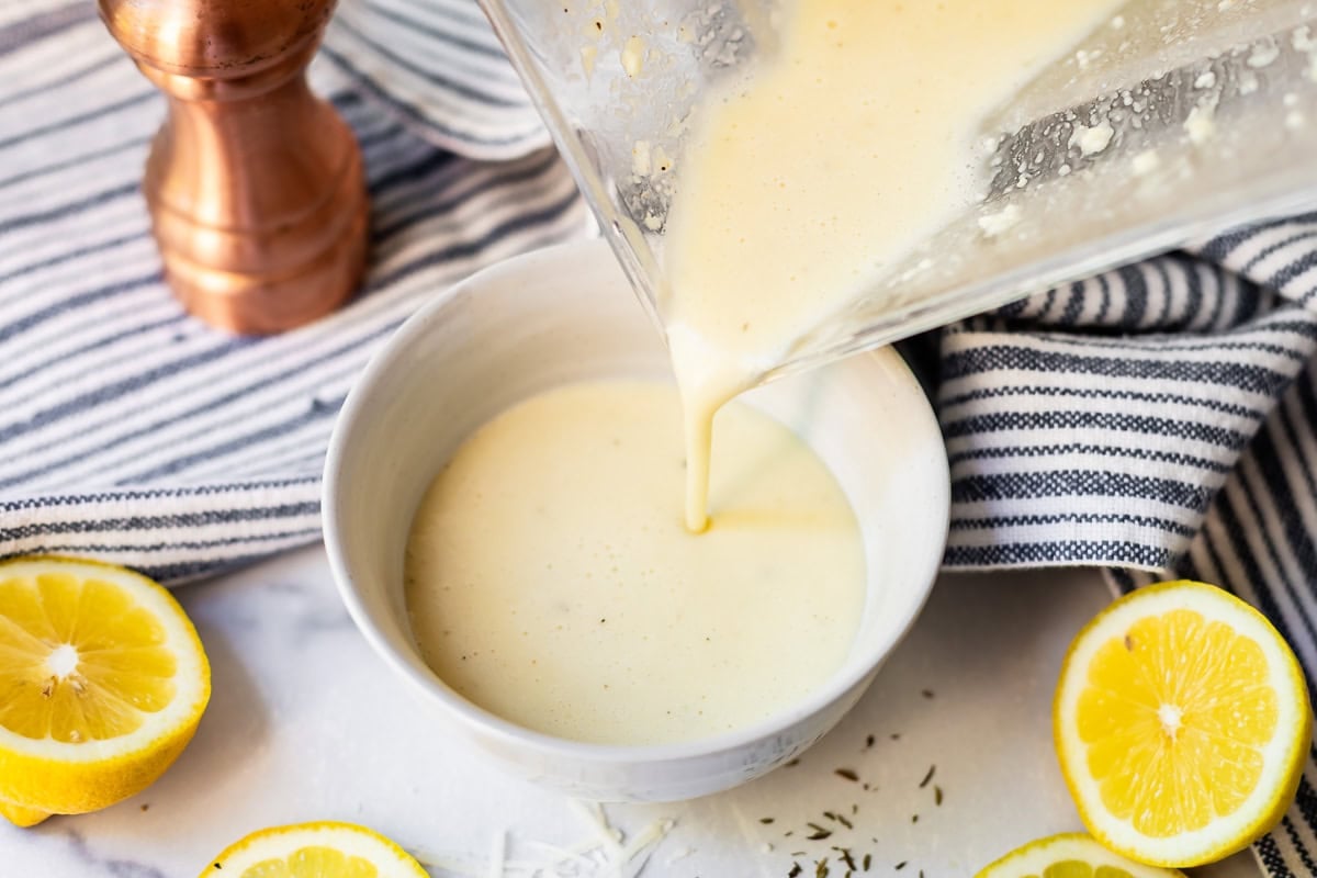 A creamy yellow sauce is being poured from a blender into a white bowl, surrounded by lemon halves, a striped towel, a pepper mill, and sprinkled herbs on a white surface.