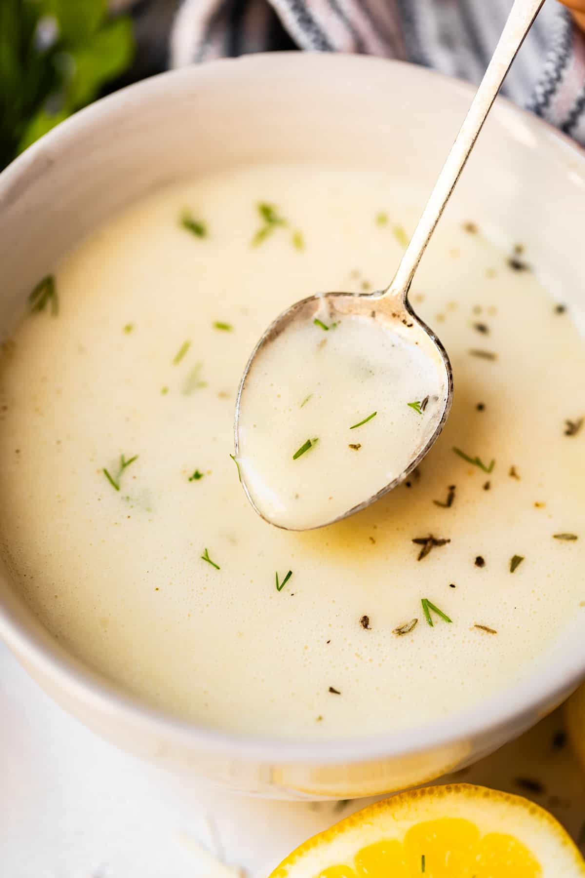 A close-up of a bowl of creamy soup, garnished with chopped herbs, with a spoon dipped inside. A lemon slice is visible in the foreground. The soup appears smooth and pale, possibly a lemon or herb-based soup.