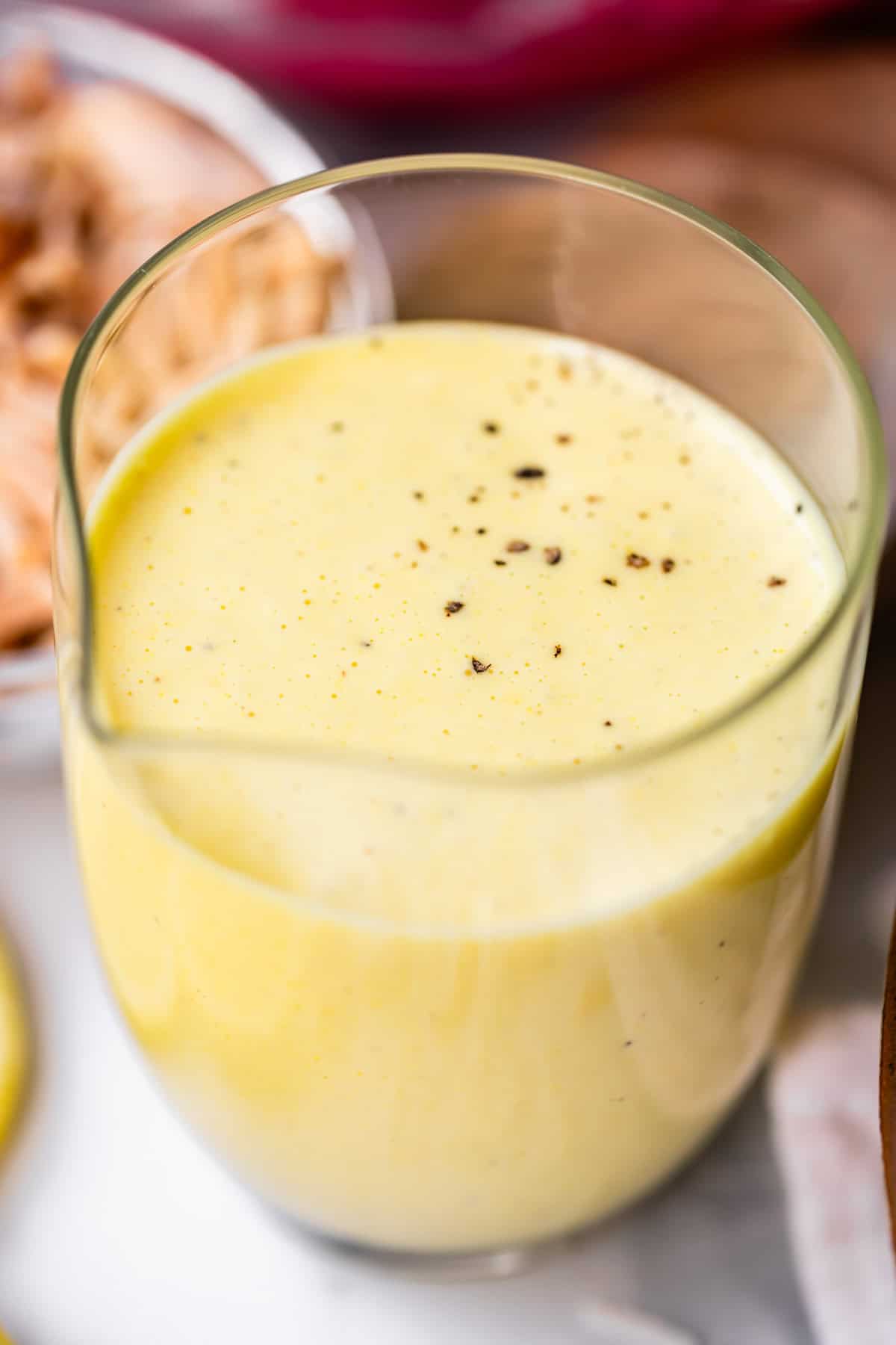 A clear glass filled with creamy yellow golden milk, topped with a sprinkle of black pepper, sits on a white surface. The background shows blurred ingredients, possibly nuts or spices.