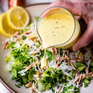 A hand pours creamy yellow dressing onto a fresh green salad topped with shredded cheese and sliced almonds. Lemon slices, a pepper grinder, and a salad bowl are visible in the background.