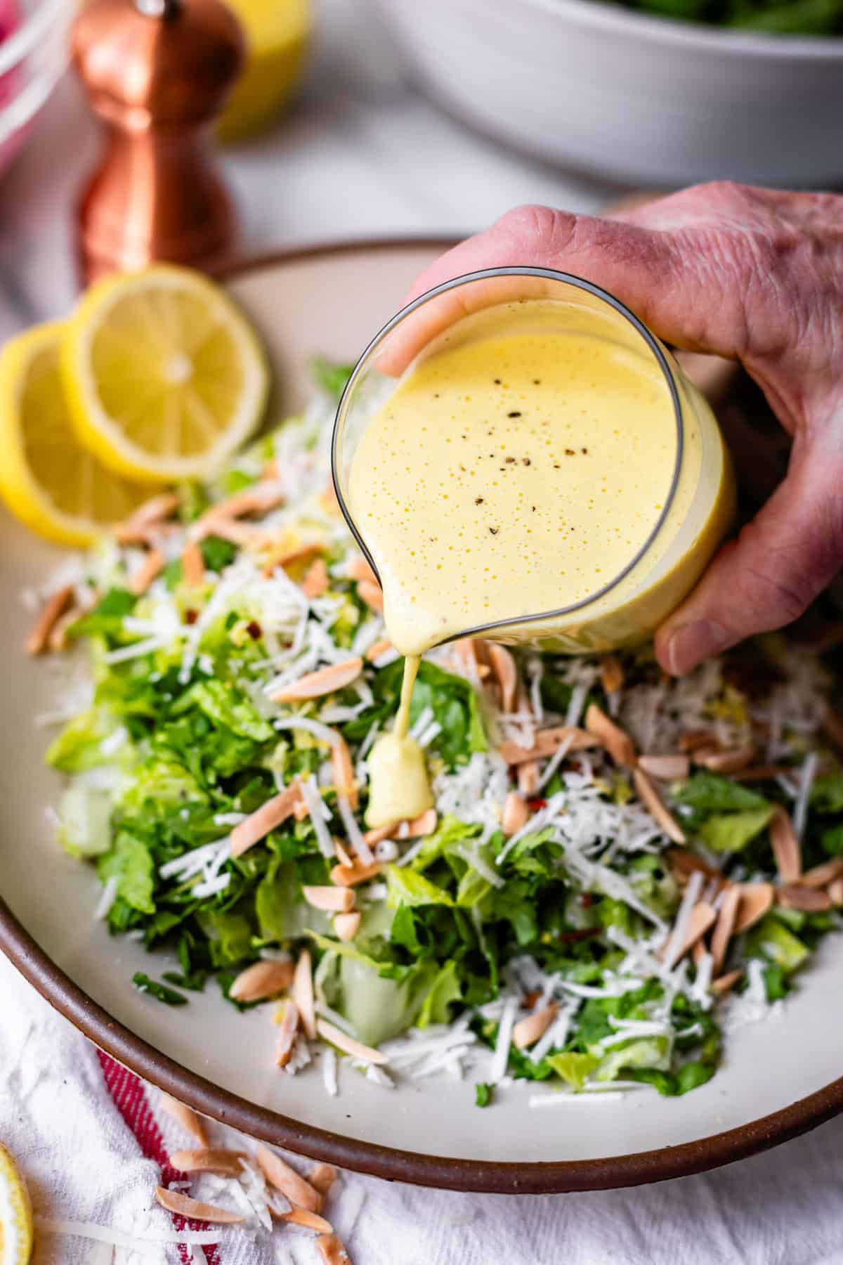 A hand pours creamy yellow dressing onto a fresh green salad topped with shredded cheese and sliced almonds. Lemon slices, a pepper grinder, and a salad bowl are visible in the background.