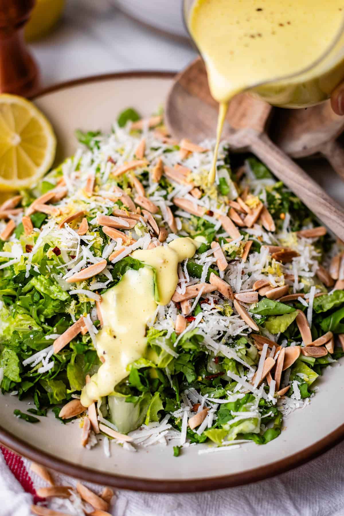 A close-up of a fresh salad topped with shredded cheese and slivered almonds, with creamy yellow dressing being poured over it. Sliced lemon and wooden salad utensils are visible on the side.