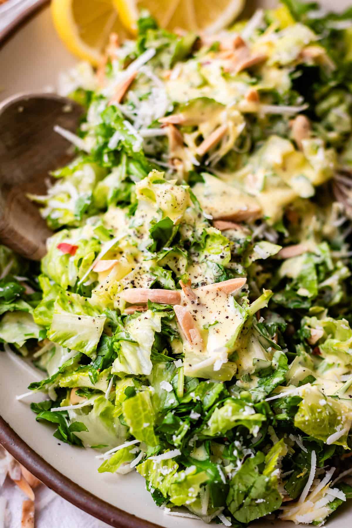 A close-up of a fresh green salad with chopped lettuce, shredded cheese, slivered almonds, creamy dressing, and cracked black pepper, garnished with lemon slices in the background.