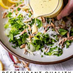 A hand pours creamy yellow lemon citrus vinaigrette over a fresh green salad topped with shredded cheese and slivered almonds on a plate. Sliced lemons and a copper pepper mill are in the background.