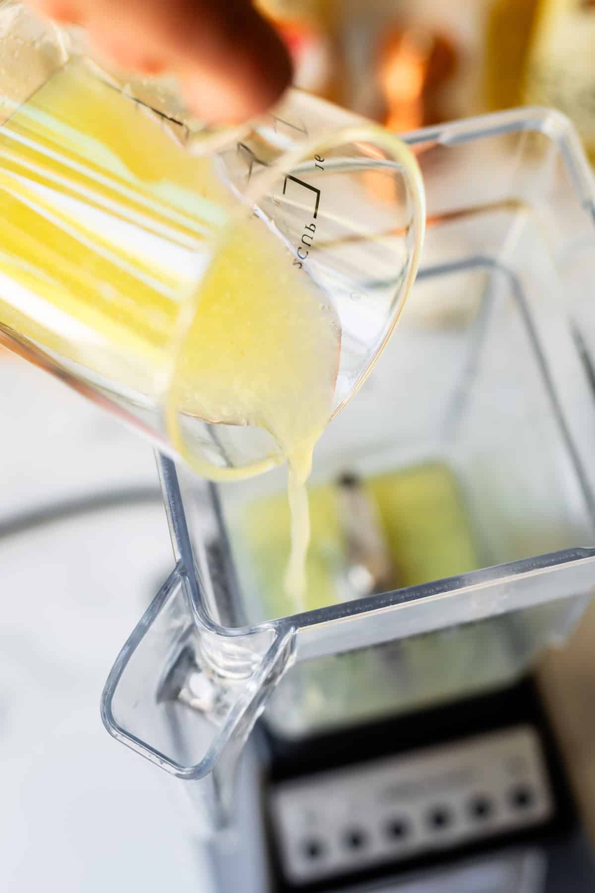 A close-up of a hand pouring a yellow liquid from a measuring cup into a blender, with the blender blades visible inside the container.