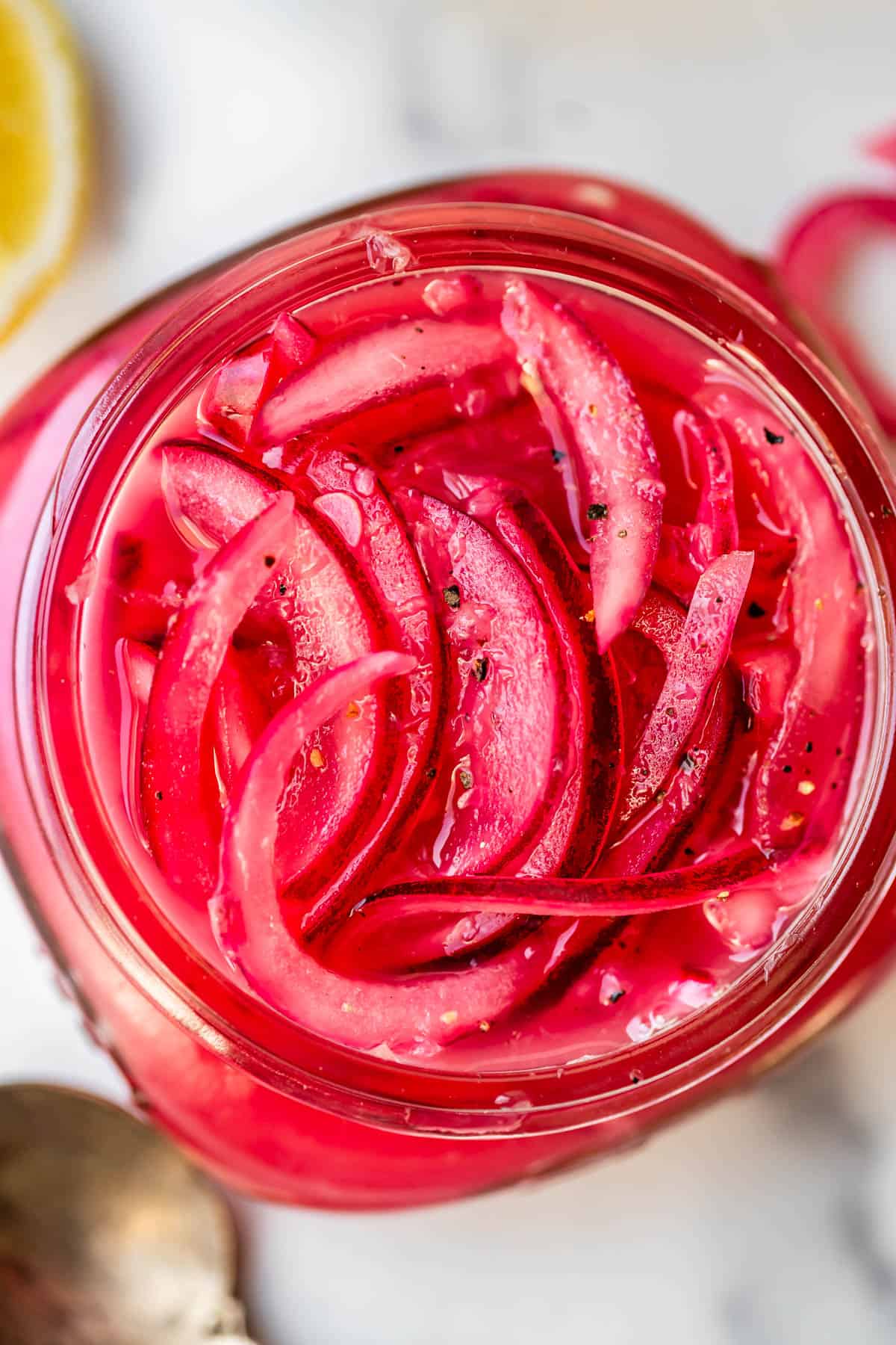 A close-up view of thinly sliced red onions pickled in a glass jar, showing vibrant pink hues and a few visible black pepper specks on top.